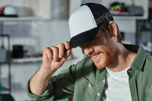 Preview: Smiling young redhead craftsman wearing snapback while working and standing in blurred print studio