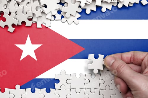 Preview: Cuba flag is depicted on a table on which the human hand folds a puzzle of white color.