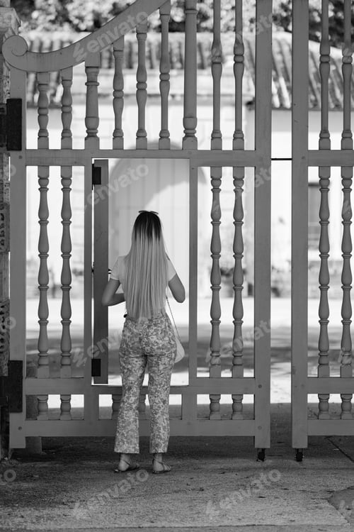 Preview: Grayscale of a young female stands among a white picket fence, opening the gate
