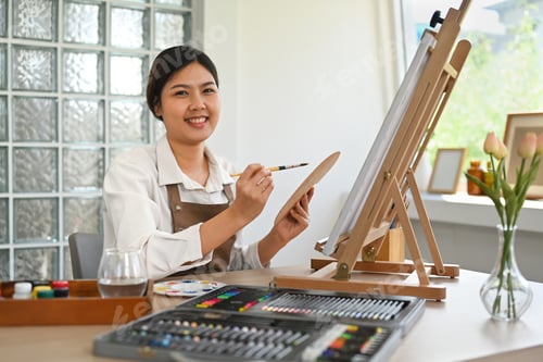Preview: Attractive female artist sitting in front of canvas and painting with watercolor.