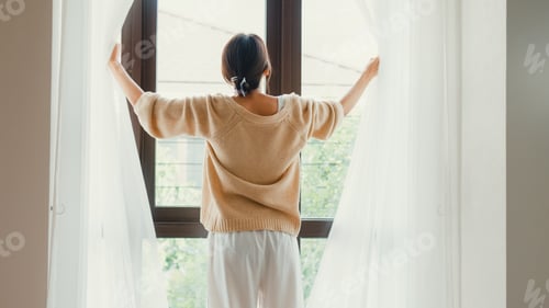 Preview: Asian girl with open white curtain on window looking outside feel fresh with peaceful morning.