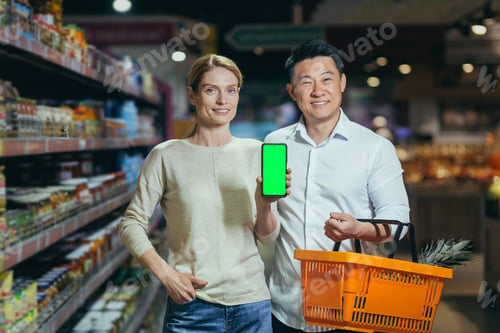 Preview: Young family diverse couple of shoppers in supermarket, smiling and looking at camera, grocery