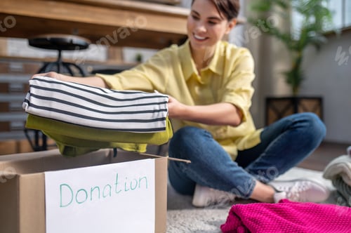Preview: Joyful girl seated on the carpet packing things for donation