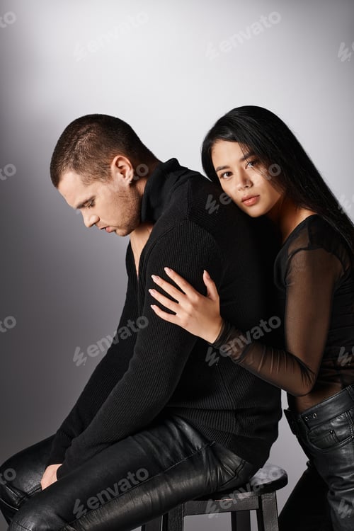 Preview: sensual asian woman embracing young upset man sitting on grey backdrop, affection and fashion