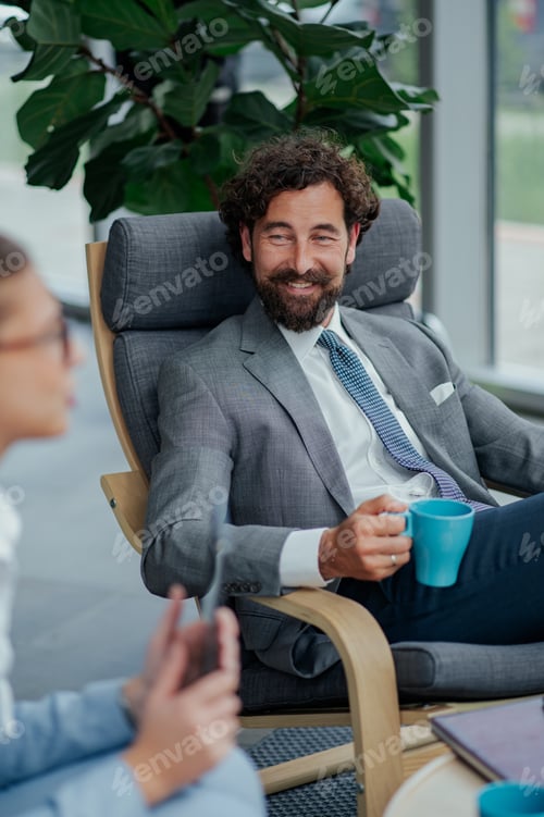 Preview: Smiling businessman relaxing on chair holding coffee mug and talking with colleague