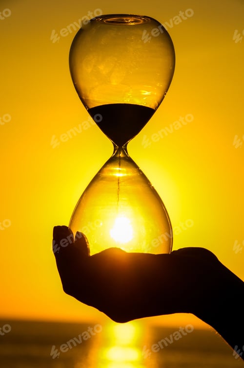 Preview: Vertical shot of a hand holding an hourglass near a golden sunset