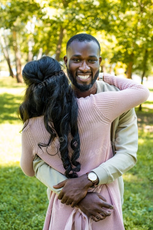 Preview: smiling african american boyfriend hugging girlfriend and looking at camera in park