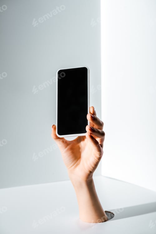 Preview: cropped image of woman holding smartphone with blank screen in hand through hole on white