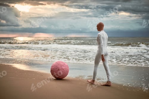 Preview: Hairless performer girl with alopecia in futuristic suit walking along seacoast with pink sphere