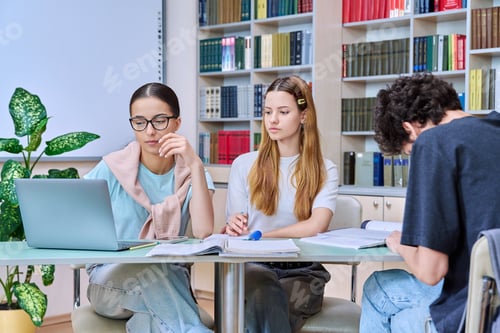 Preview: Group of students teenagers sitting together at desk in library preparing for tests exams