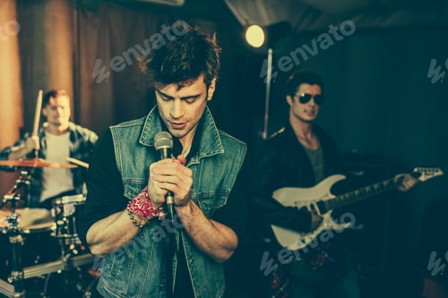 Preview: selective focus of fashionable man singing song in microphone near rock band