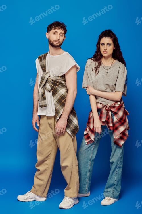 Preview: couple posing in street wear, blue backdrop, woman with bold makeup holding cap, bearded man