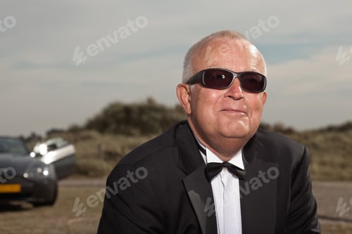 Preview: Senior Good Looking Happy Relaxed Retired Man With His Sports Car In Dune Landscape With Cloudy