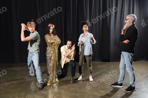 Preview: grey haired art director looking at multiracial woman in queen costume standing with crossed arms