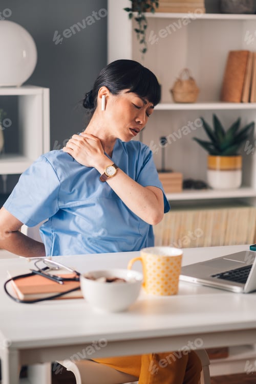 Preview: Asian woman ealthcare professional in scrubs massages neck at work desk