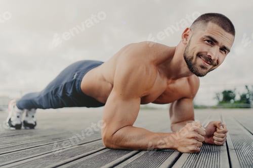 Preview: Pleased muscular bearded young man stands in plank pose, makes sport training outdoors