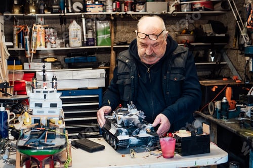 Preview: Senior man, pensioner holding made handicraft scale model of a tank on the table ih his garage