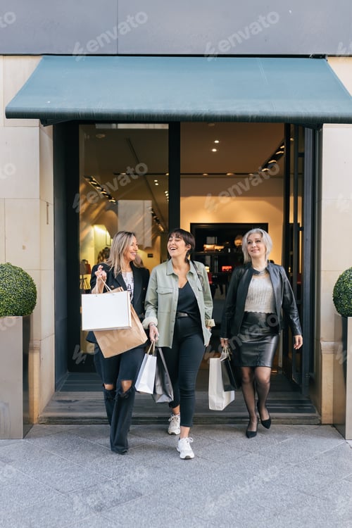 Preview: Happy ethnic women friends coming out of shopping center with paper bags in daylight
