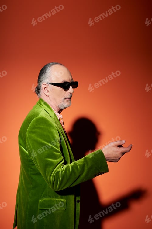 Preview: elderly and grey haired man pointing with hand and looking away on red and orange background