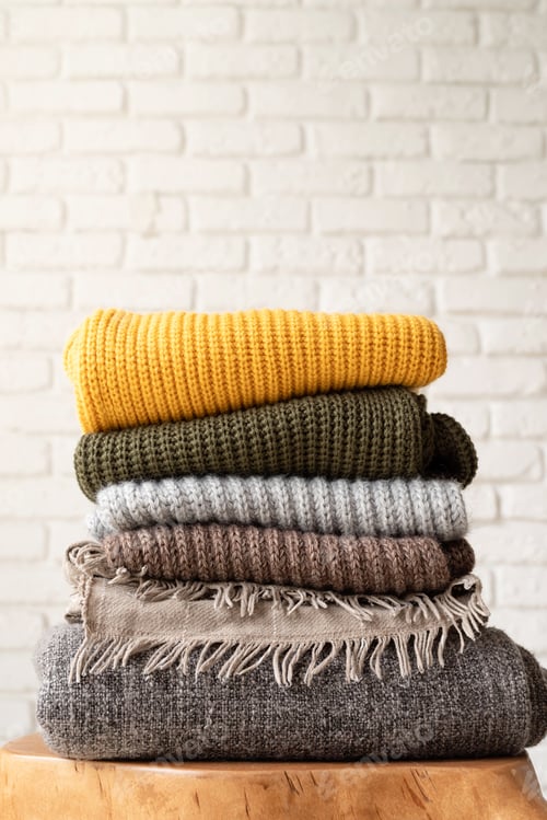 Preview: Stack of autumn warm sweaters on modern coffee table, white brick wall background