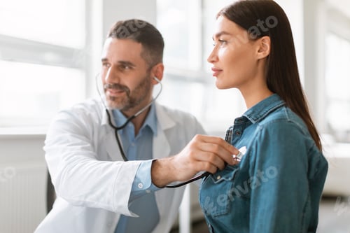 Preview: Caring male doctor using phonendoscope, examining young female patient heart rate at consultation in