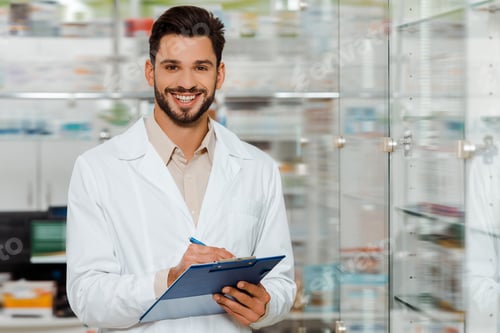 Preview: Handsome pharmacist with clipboard smiling at camera by drugstore showcase