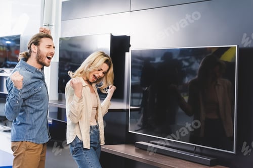 Preview: happy boyfriend and girlfriend showing yes gesture near tv in home appliance store