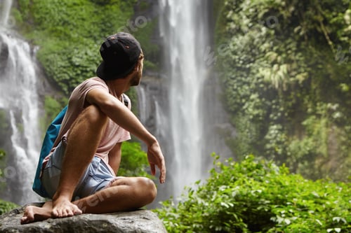 Preview: People, lifestyle and adventure concept. Stylish young man wearing cap backwards sitting on big rock