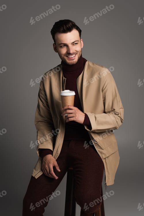 Preview: Smiling man in autumn outfit holding coffee to go while sitting on chair isolated on grey
