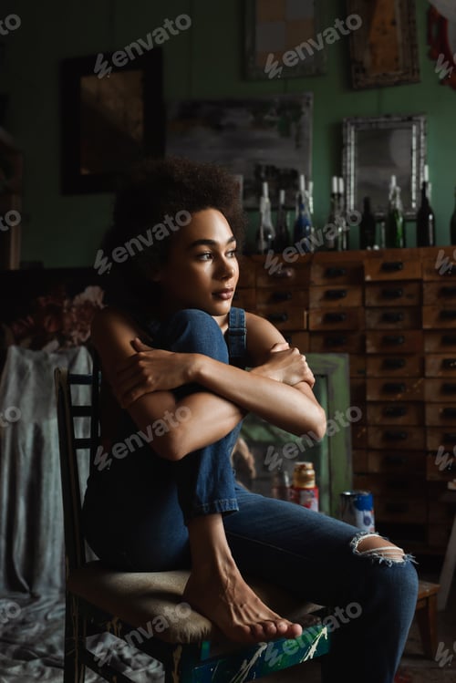 Preview: pretty and thoughtful african american woman in overalls sitting with crossed arms in art studio