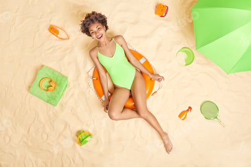 Preview: Positive curly haired Afro American woman in green swimwear poses on inflated lifebuoy smiles pleasa