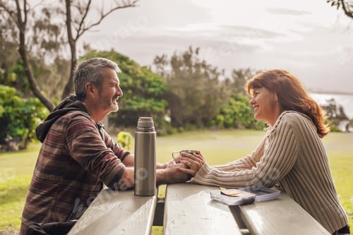 Preview: Middle-aged couple sitting on the benches with table in park and drinking tea.