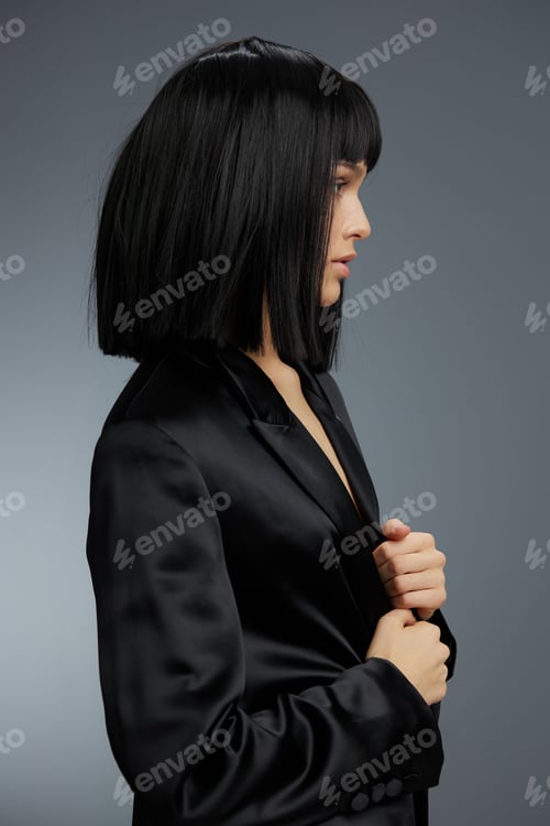Preview: Young woman with black bob hairstyle wearing a black suit and blazer standing against a gray