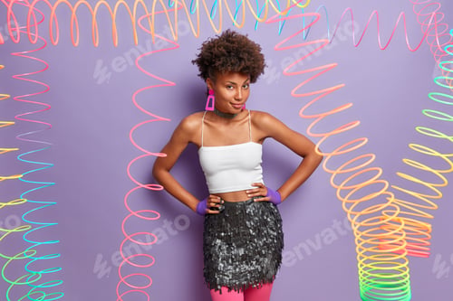 Preview: Self assured serious dark skinned woman keeps hands on hips, dressed in white top and skirt, looks m
