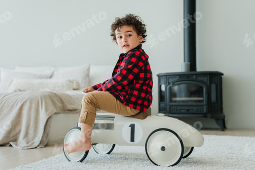 Preview: Curly caucasian little boy in plaid shirt, pants sitting on toy car at bedroom looks at camera