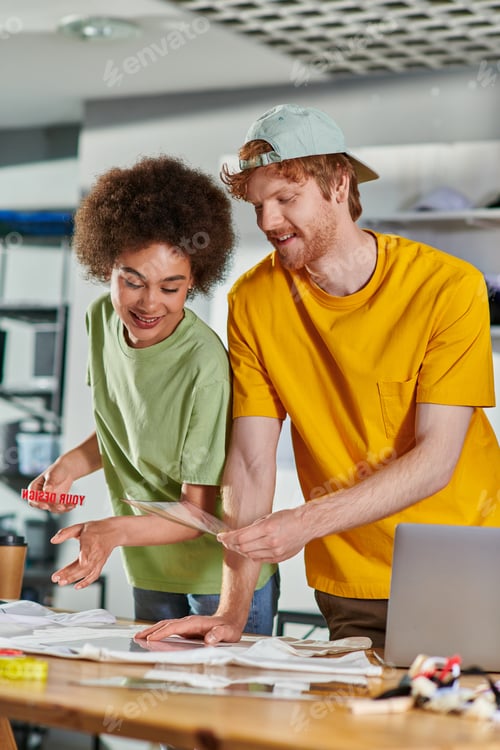 Preview: Smiling craftsman holding printing layer near african american colleague, clothes and laptop