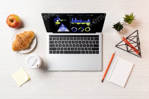 Preview: top view of laptop with business charts, croissant, apple and paper cup at workplace