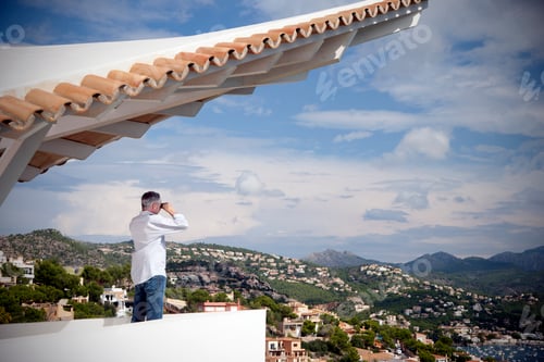 Preview: Spain, Mallorca, man standing on terrace of a house looking at view with binocular