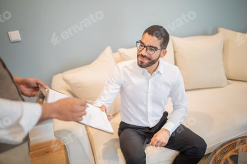 Preview: A man in a white shirt is sitting on a couch and receiving a document