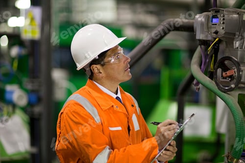 Preview: Industrial worker in orange safety gear inspecting machinery with concentration.
