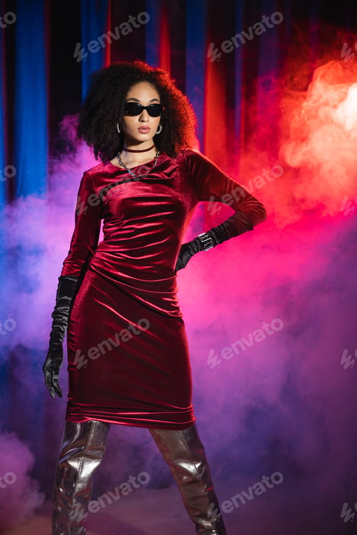 Preview: Stylish african american woman in sunglasses and velvet dress on red background with blue light