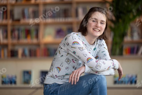 Preview: Smiling person in a casual sweater and jeans sitting in a library