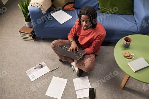Preview: Young Black Man Typing on Laptop Surrounded by Papers in Living Room