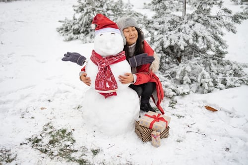 Preview: Asian girl building snowman in snowy park. Woman embracing snowman wearing hat and scarf. Active out
