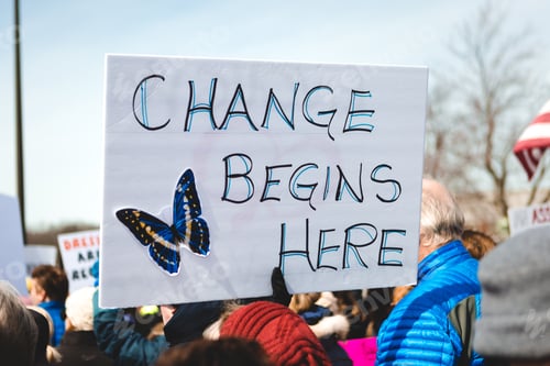 Preview: Activist sign with butterfly reads “change begins here” held by a gloved hand in a peaceful protest