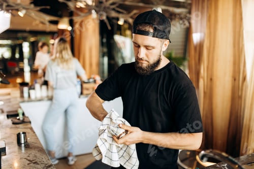 Preview: A young stylish man with beard,wearing casual clothes, wipes a glass in a modern coffee shop