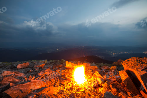 Preview: Beautiful summer night in mountain. Bright fire burns on rocky mountain top under blue cloudy sky.