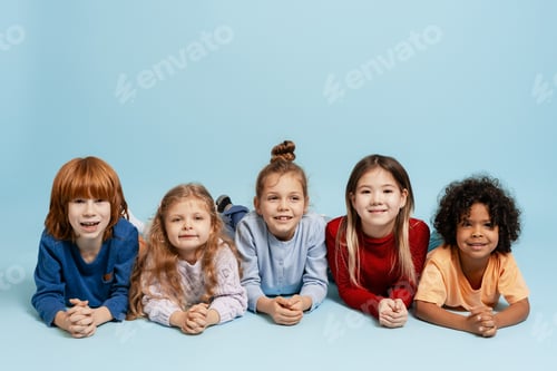 Preview: Diverse group of smiling children lying together on light blue background, looking at camera