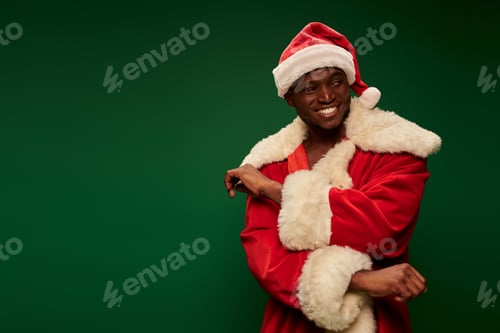 Preview: joyful african american guy in festive santa claus costume looking away on green backdrop