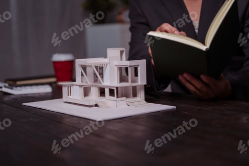 Preview: A businessman sits at a table at night, holding a model of a two-story white house. He reviews